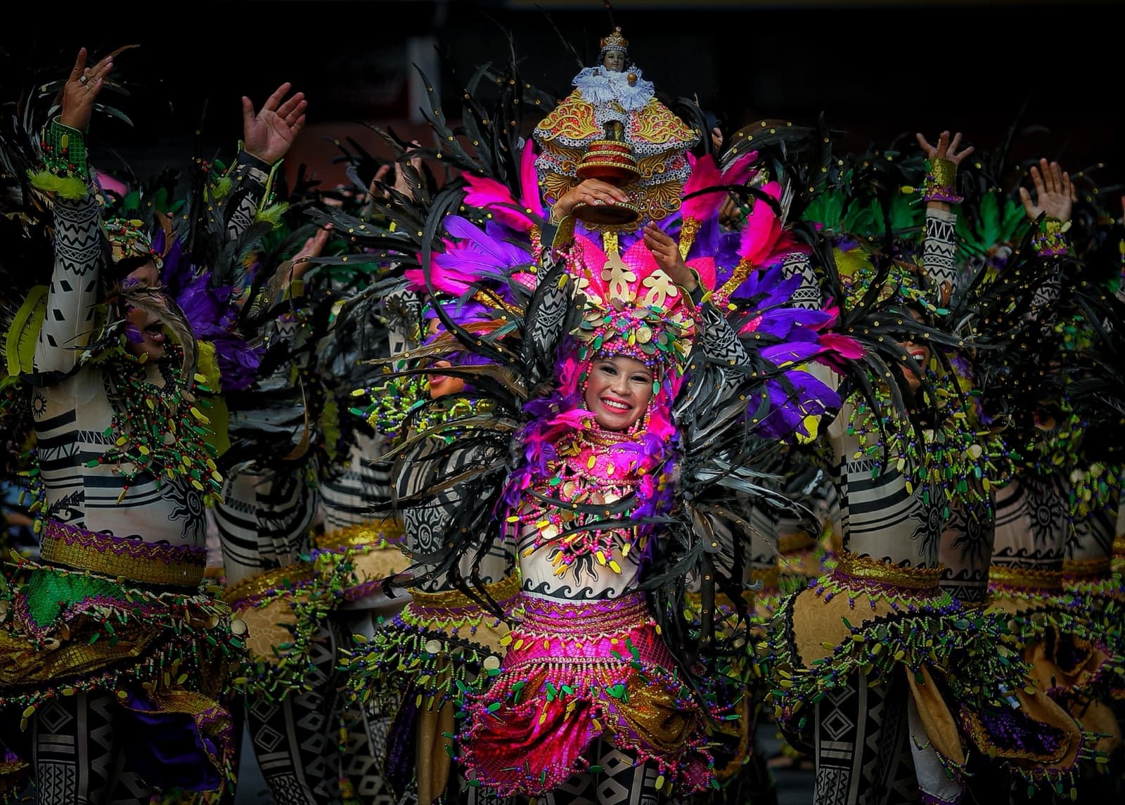 A Sinulog Festival dancer in a brilliant costume of pink, purple and green feathers, smiling with arms raised