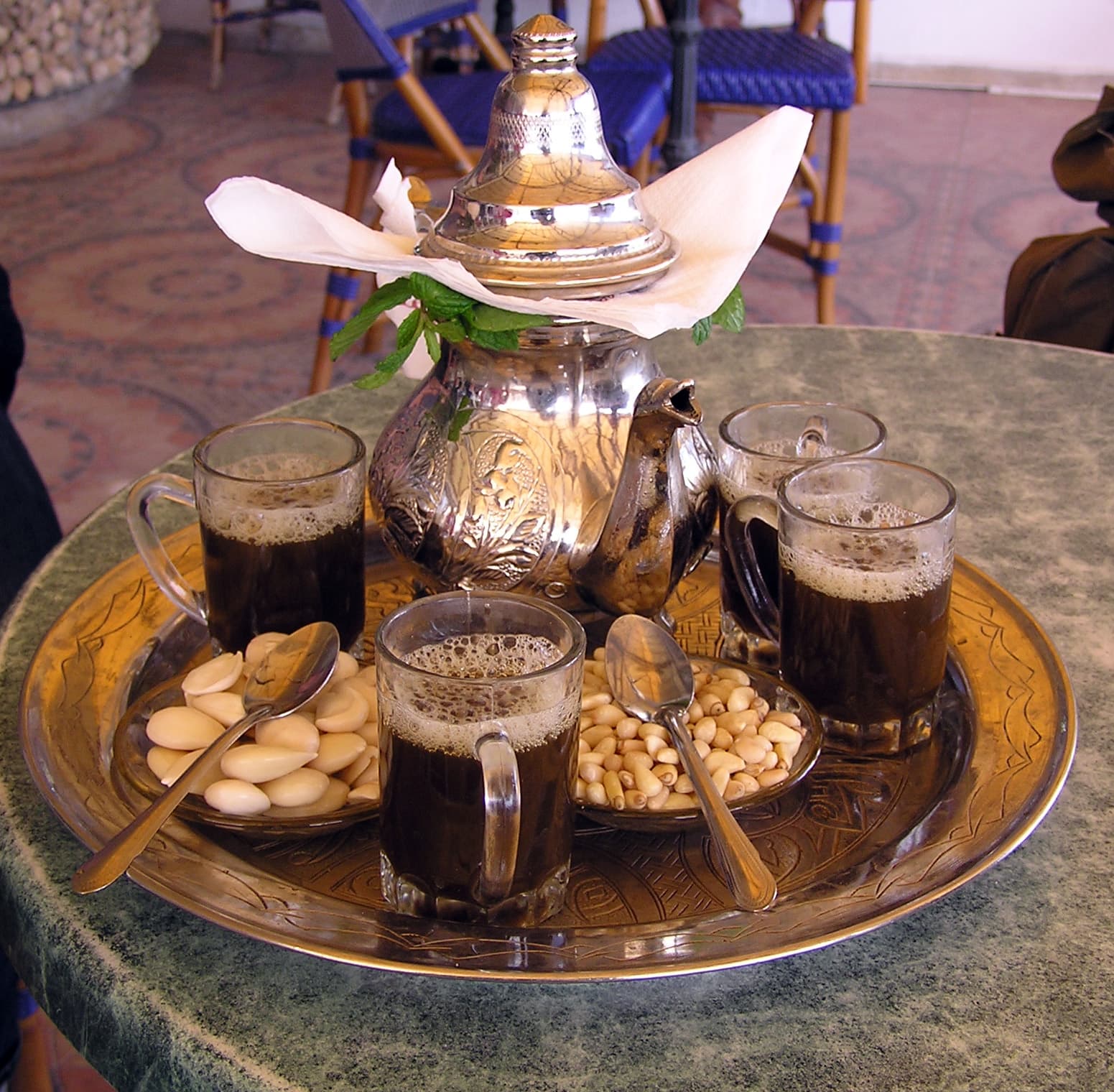 A small glass of bright green mint tea being poured from a silver teapot held high above